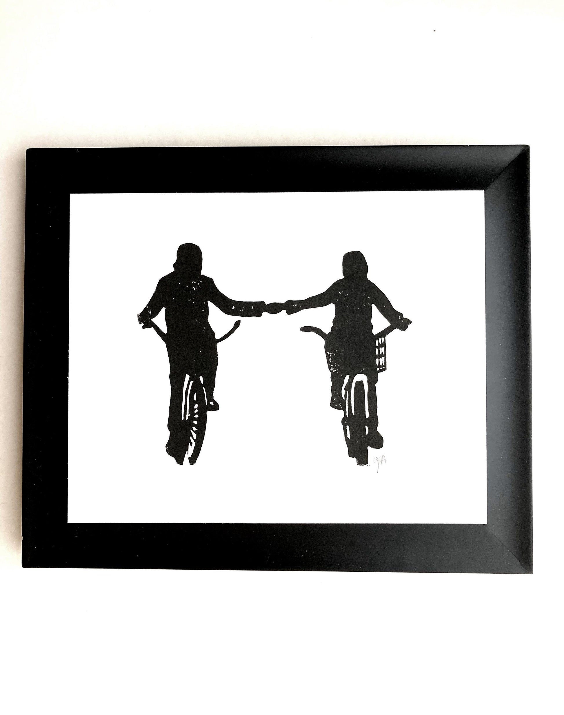 Framed black and white silhouette artwork of two people on bicycles holding hands.